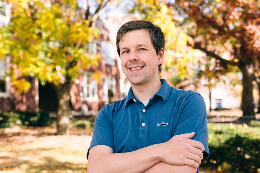 David Gleich standing with arms crossed in a blue polo shirt, autumn trees and a brick building behind him